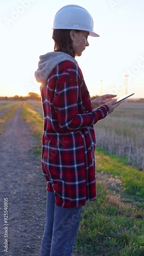 Woman engineer wearing a white protective helmet is taking notes with a clipboard in a field with wind turbines, as the sun sets. Clean energy and engineering concept