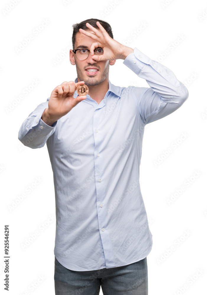 Handsome young man holding bitcoin with happy face smiling doing ok sign with hand on eye looking through fingers Handsome young man holding bitcoin with happy face smiling doing ok sign with hand on eye looking through fingers