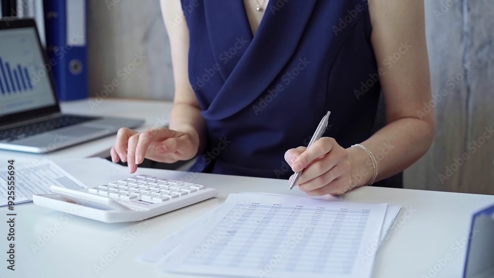 Fototapeta premium Businesswoman with dark blue dress is using white calculator and taking notes in her office. Taxes and audit in business