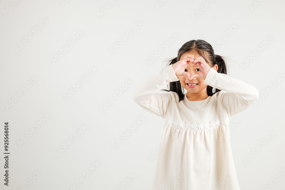 A smiling Asian young kid girl shows confidence, creating a heart shape with fingers and hands on white background. Portrait of kindergarten child sending love for Valentine's Day A smiling Asian young kid girl shows confidence, creating a heart shape with fingers and hands on white background. Portrait of kindergarten child sending love for Valentine's Day