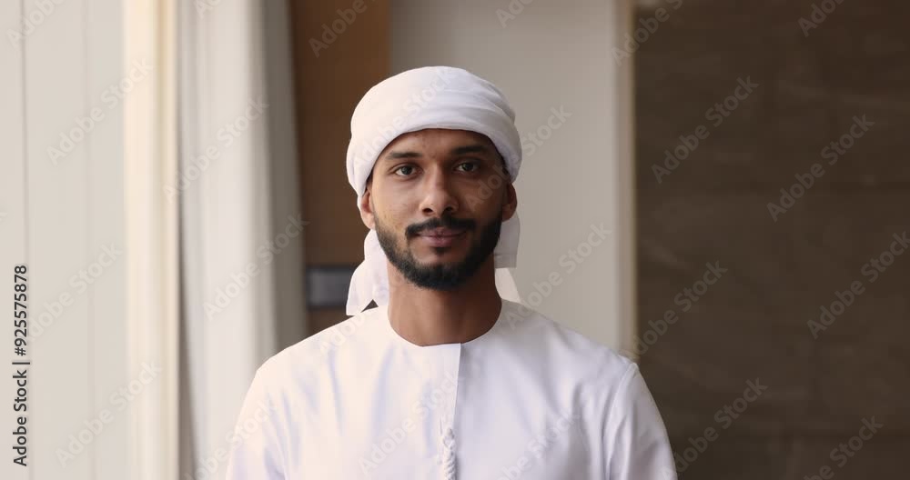 Serious handsome young Arabic business owner man in white Muslim clothes posing in office for front portrait, getting happy, looking at camera. Successful Arab businessman, boss, leader head shot