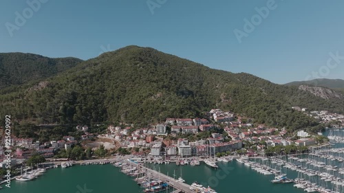 A scenic view of a marina nestled at the base of a lush green mountain, with boats and yachts docked in the clear blue water under a bright sky. photo