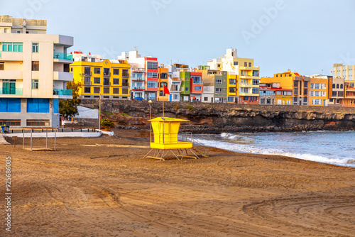 Playa de la Garita in Gran Canaria, Canary Islands, Spain. Yellow lifeguard stand sits on a sandy beach next to ocean. Beach scene featuring a lifeguard stand with buildings visible in the background photo