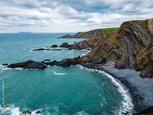 Scenic North Devon coastline with shingle beach and beautiful rock formations. Aerial view at Eye Cove on a cloudy day. Rocks covered with moss. Lundy island at distance. Ocean waves.