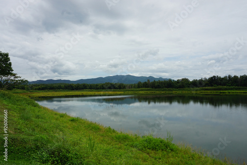 A serene landscape photo showcasing a lake surrounded by lush greenery and mountains in the distance. The sky is overcast with soft, white clouds. The lake is calm and reflective, mirroring the beauty