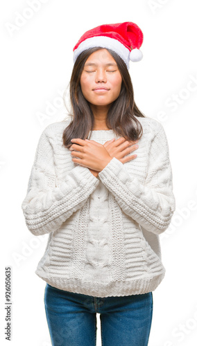 Young asian woman wearing christmas hat over isolated background smiling with hands on chest with closed eyes and grateful gesture on face. Health concept.