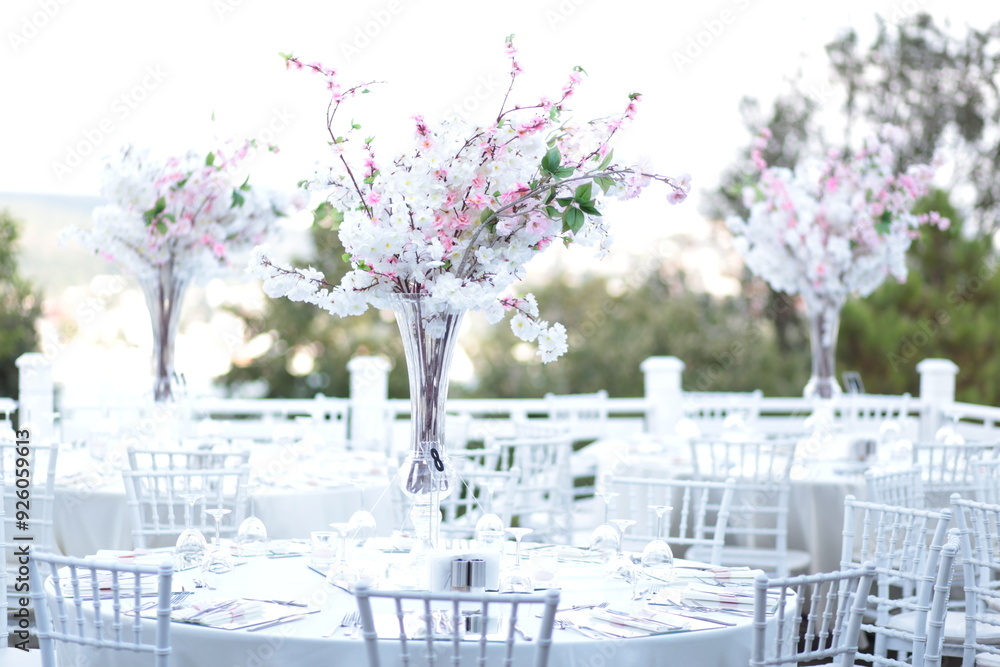 A romantic and elegant outdoor wedding reception with round tables and tall floral centerpieces. The flowers are a mix of pink and white, creating a beautiful and dreamy setting