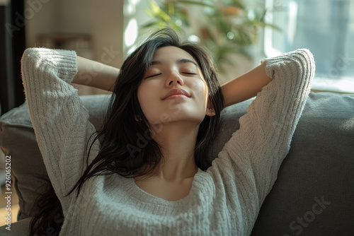 Cozy living room scene with a young Asian woman relaxing on the sofa, hands behind her head, eyes closed, wearing casual clothes, presented in a calm and serene photorealistic style.