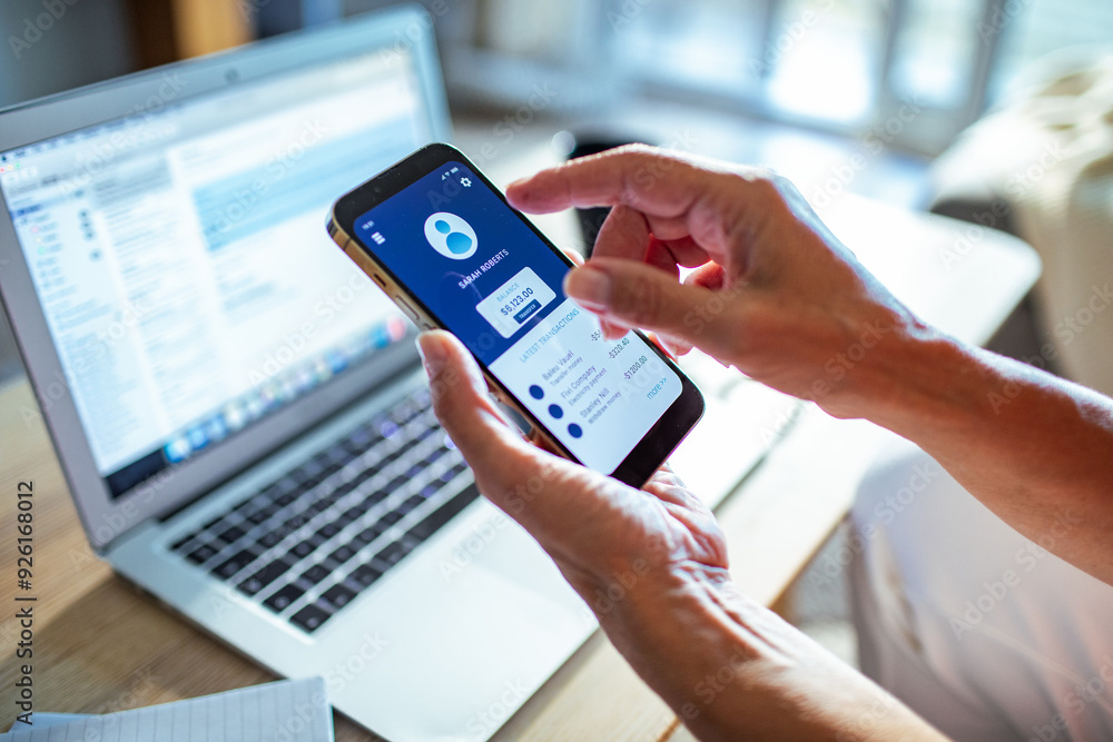 Senior woman checking bank account on smartphone with laptop in the background at home Senior woman checking bank account on smartphone with laptop in the background at home