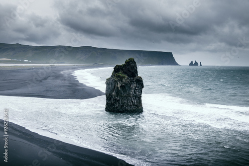 Einsamer Basaltfelsen am schwarzen Sandstrand von Dyrhólaey in Island, umgeben von tosenden Wellen des Atlantiks, mit der markanten Küstenlinie von Reynisdrangar im Hintergrund
