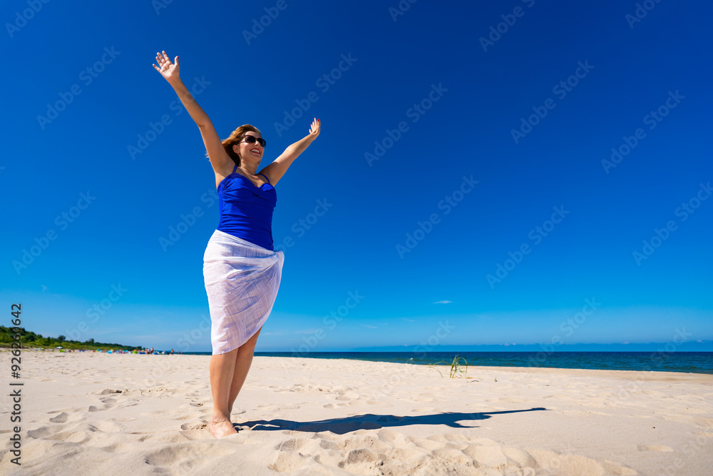 Summer vacation. Beautiful young woman in one-piece blue swimsuit and white scarf and sunglasses standing with her arms raised up on sandy beach on beautiful sunny day. Freedom and power. Summer vacation. Beautiful young woman in one-piece blue swimsuit and white scarf and sunglasses standing with her arms raised up on sandy beach on beautiful sunny day. Freedom and power.