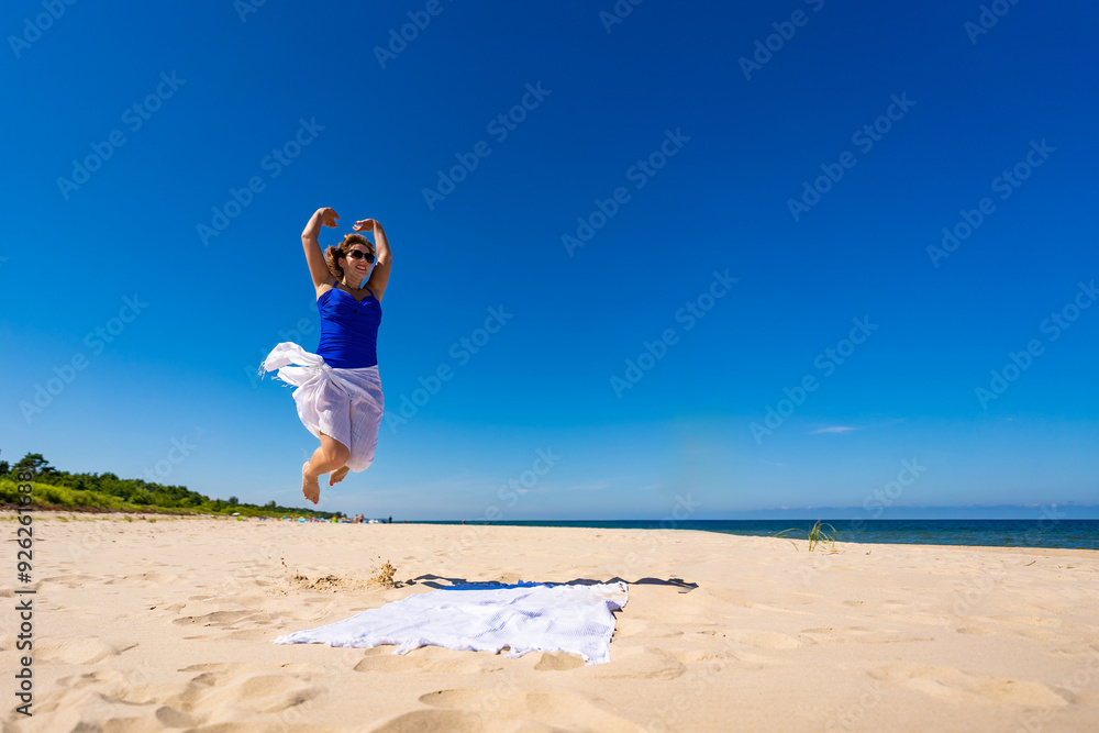 Φωτογραφία Young woman in blue swimsuit, white pareo and sunglasses jumping high up on white sand on beautiful beach against blue sky on sunny day