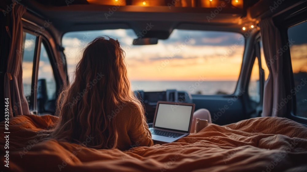 Fototapeta premium A woman relaxes in a warm camper van, illuminated by string lights, as she works on her laptop while watching a breathtaking sunset