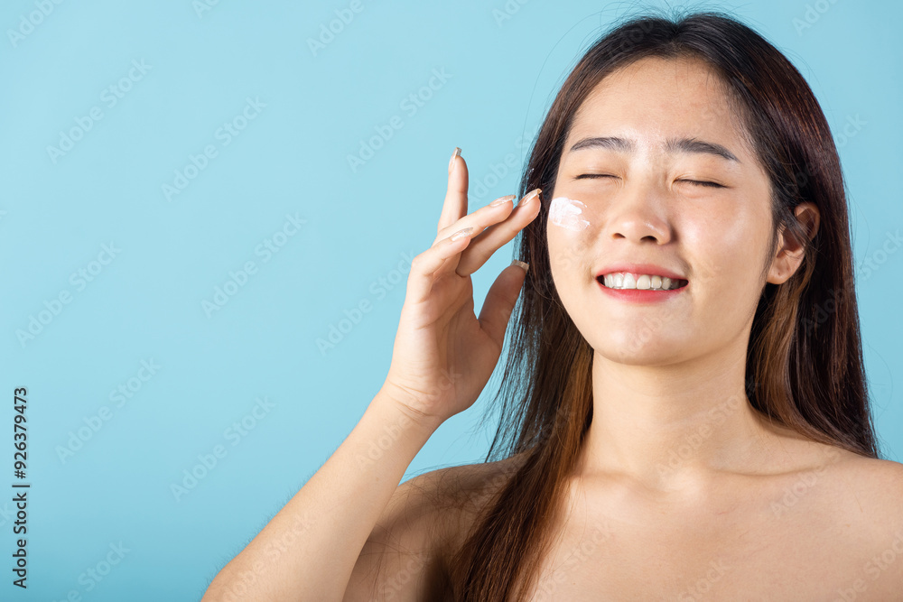 Happy beauty portrait woman gets cream on her cheek, Beautiful young female smiling applying cosmetic cream on her face isolated on blue background, health clean skin, Skin care and beauty concept Happy beauty portrait woman gets cream on her cheek, Beautiful young female smiling applying cosmetic cream on her face isolated on blue background, health clean skin, Skin care and beauty concept