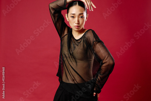 Fashion asian woman in sheer top and skirt posing against red background with hands on hips and head tilted to the side