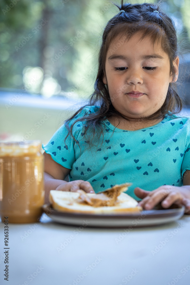 A young girl is sitting at a table spreading the peanut butter on a slice of bread. A young girl is sitting at a table spreading the peanut butter on a slice of bread.