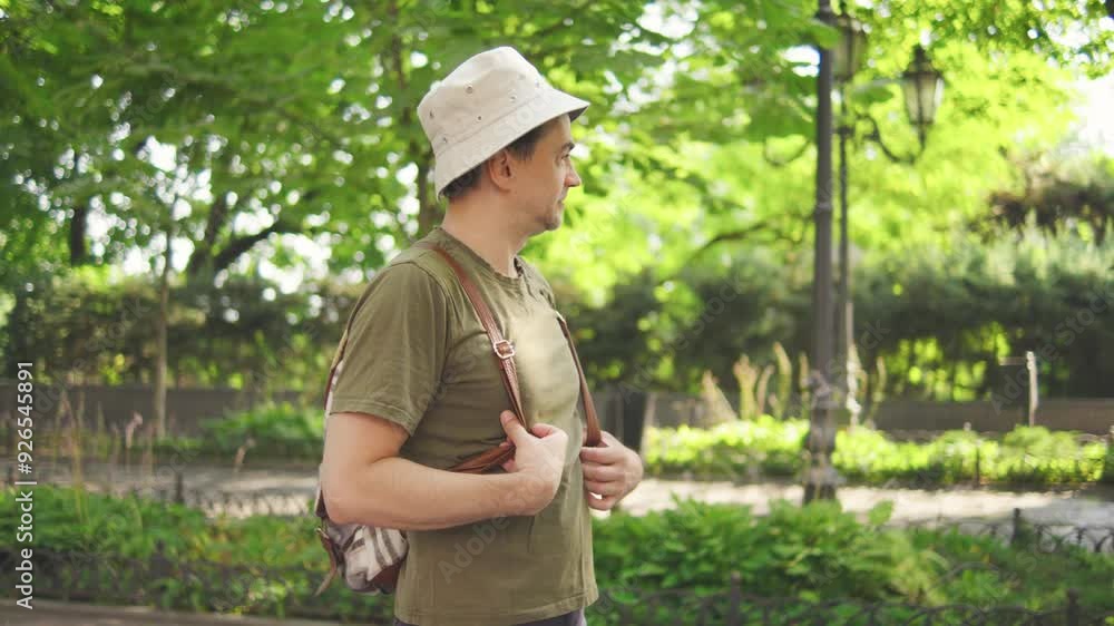 An adult tourist wearing a Panama hat and carrying a backpack enjoys a beautiful, lush park, the freshness of nature and the joy of exploring a new city An adult tourist wearing a Panama hat and carrying a backpack enjoys a beautiful, lush park, the freshness of nature and the joy of exploring a new city