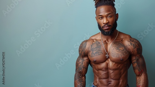 A muscular man with tattoos poses confidently against a solid blue background in a studio setting