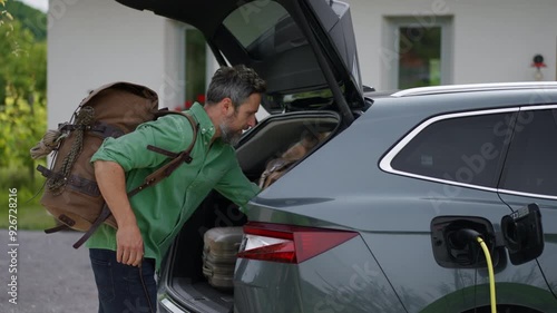 Father with son loading luggage into trunk of electric car with charger in charging port. Travelling on family road trip or vacation by electric vehicle.
