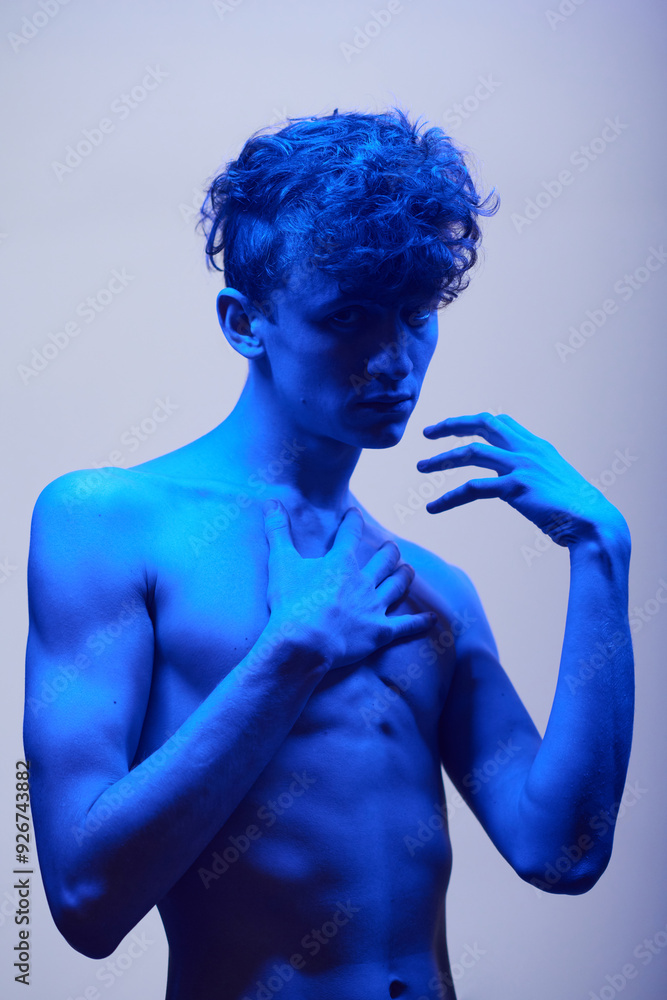 Young man striking a thoughtful pose with one hand on his chest and dramatic blue lighting casting shadows on his features Young man striking a thoughtful pose with one hand on his chest and dramatic blue lighting casting shadows on his features