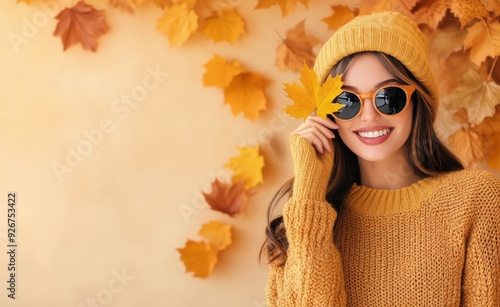Young woman wearing yellow sweater and hat holds autumn leaf against a yellow...