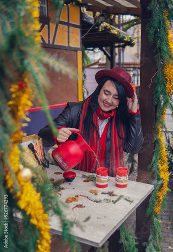 A young European woman in a red hat and scarf pours tea from a red thermos into cups at a Christmas market at a table among New Year's decorations