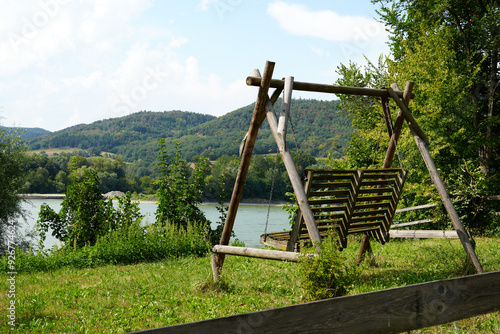 A wooden swing in a tranquil Austrian countryside, set against lush green grass, a clear sky, trees, and a gently flowing river photo