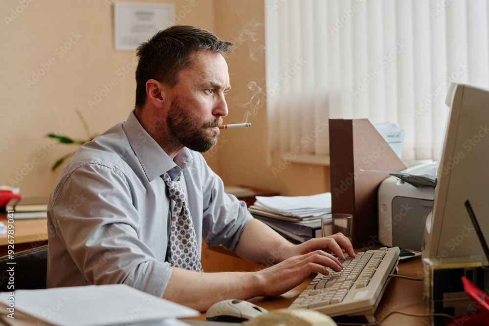 Fototapeta premium Man sitting at desk in office, smoking cigarette while typing on keyboard. Papers and office supplies scattered around giving busy work environment impression