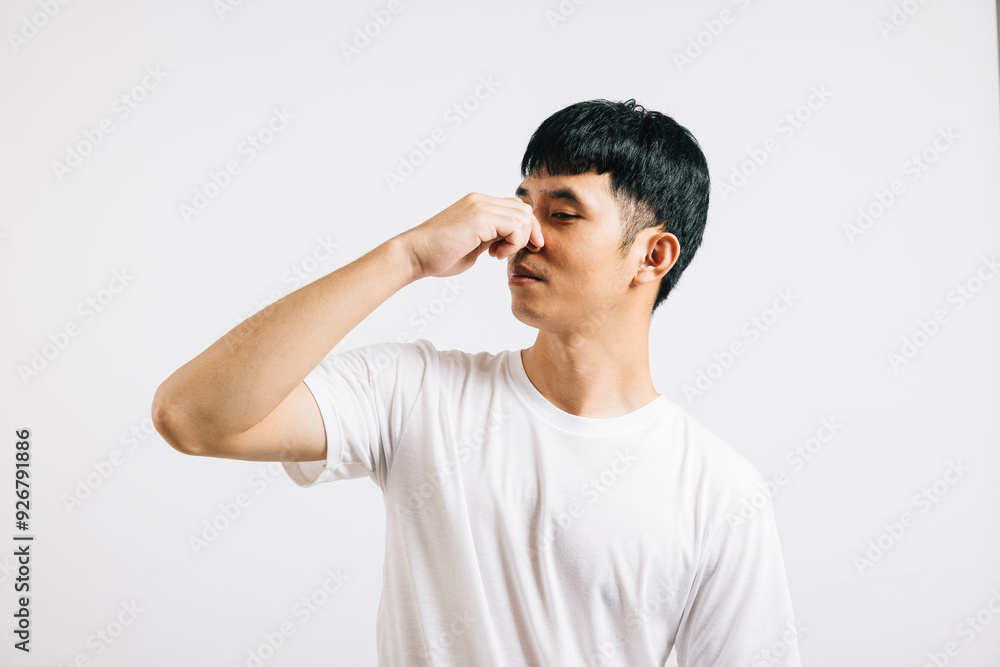 An Asian young man wears an unhappy, disgusted expression, pinching his nose due to a strong and smelly odor. Studio shot isolated on a white background, illustrating a bad smell situation. An Asian young man wears an unhappy, disgusted expression, pinching his nose due to a strong and smelly odor. Studio shot isolated on a white background, illustrating a bad smell situation.