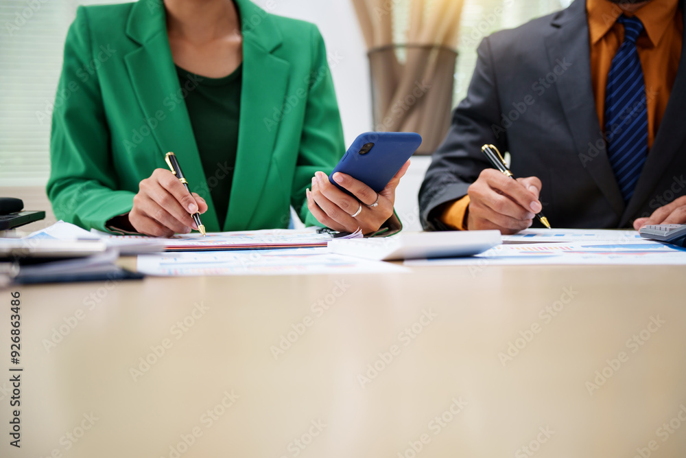 Fototapeta premium Asian business team engages in a collaborative meeting, financial charts at a desk. Dressed in formal suits, they brainstorm creative ideas to enhance company performance and team cohesion.