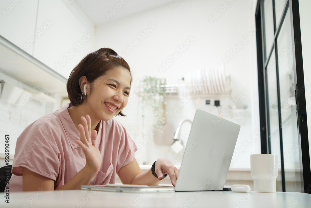 Confident businesswoman in pink shirt smiling and waving during a video call on her laptop, wearing wireless earphones, seated at a desk in a modern kitchen. Confident businesswoman in pink shirt smiling and waving during a video call on her laptop, wearing wireless earphones, seated at a desk in a modern kitchen.