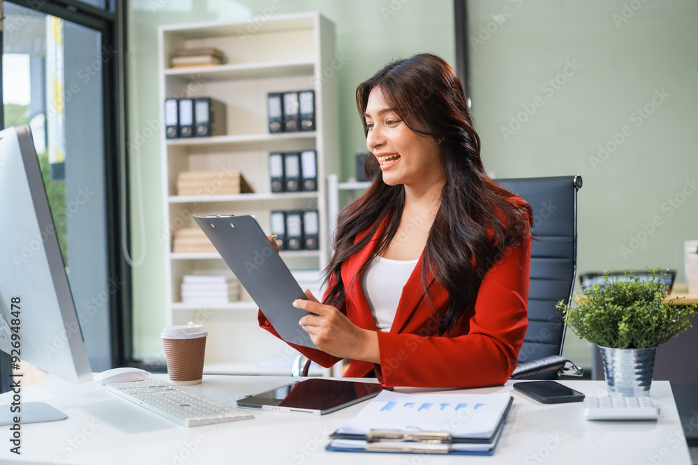 An Asian businesswoman sits at her desk, working efficiently on a computer and tablet. She smiles confidently, displaying her expertise as a financial analyst. Her work brings her happiness An Asian businesswoman sits at her desk, working efficiently on a computer and tablet. She smiles confidently, displaying her expertise as a financial analyst. Her work brings her happiness