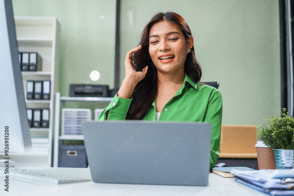 An Asian businesswoman sits at the table in her office, working as an accountant. She uses a computer with a smile, feeling happy and content with her work. An Asian businesswoman sits at the table in her office, working as an accountant. She uses a computer with a smile, feeling happy and content with her work.