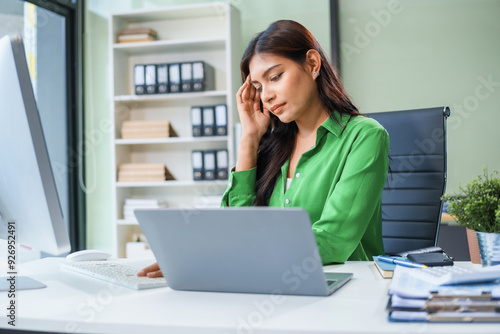 An Asian businesswoman sits at her office table, stressed and with a headache from hard work. Prolonged sitting leads to muscle weakness and the negative effects of office syndrome.