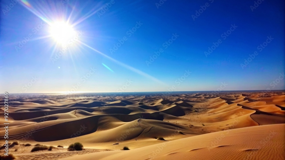 Naklejka premium A vast, golden desert stretches out under a clear blue sky, with a bright sun casting long shadows on the sand dunes. The image evokes feelings of heat, vastness, and the beauty of nature.