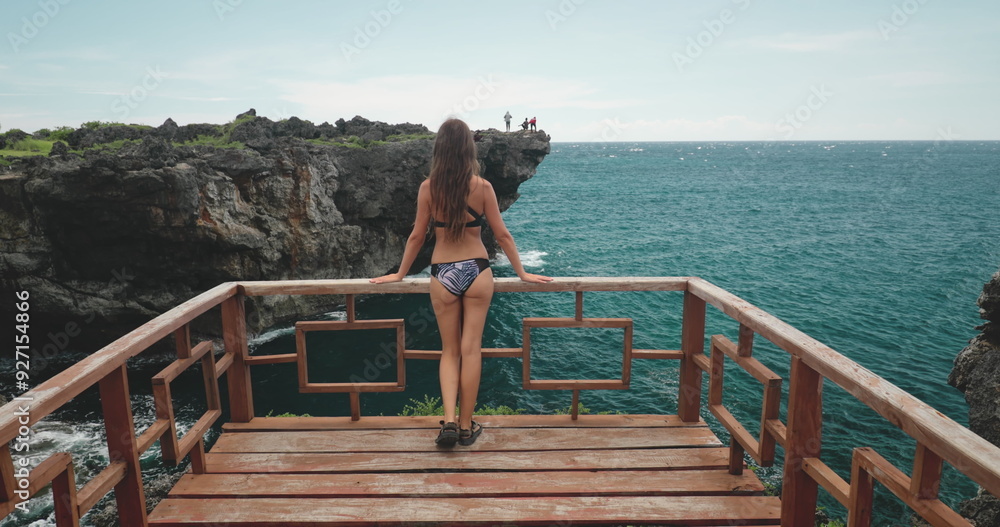 Long hair brunette in bikini walking wooden balcony with awesome views of ocean rock cliff. Tourist beautiful woman relax on seashore cliff. Travel, tourism, holiday. Back view, Long hair brunette in bikini walking wooden balcony with awesome views of ocean rock cliff. Tourist beautiful woman relax on seashore cliff. Travel, tourism, holiday. Back view,