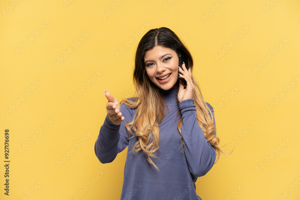 Young Russian girl using mobile phone isolated on yellow background shaking hands for closing a good deal Young Russian girl using mobile phone isolated on yellow background shaking hands for closing a good deal