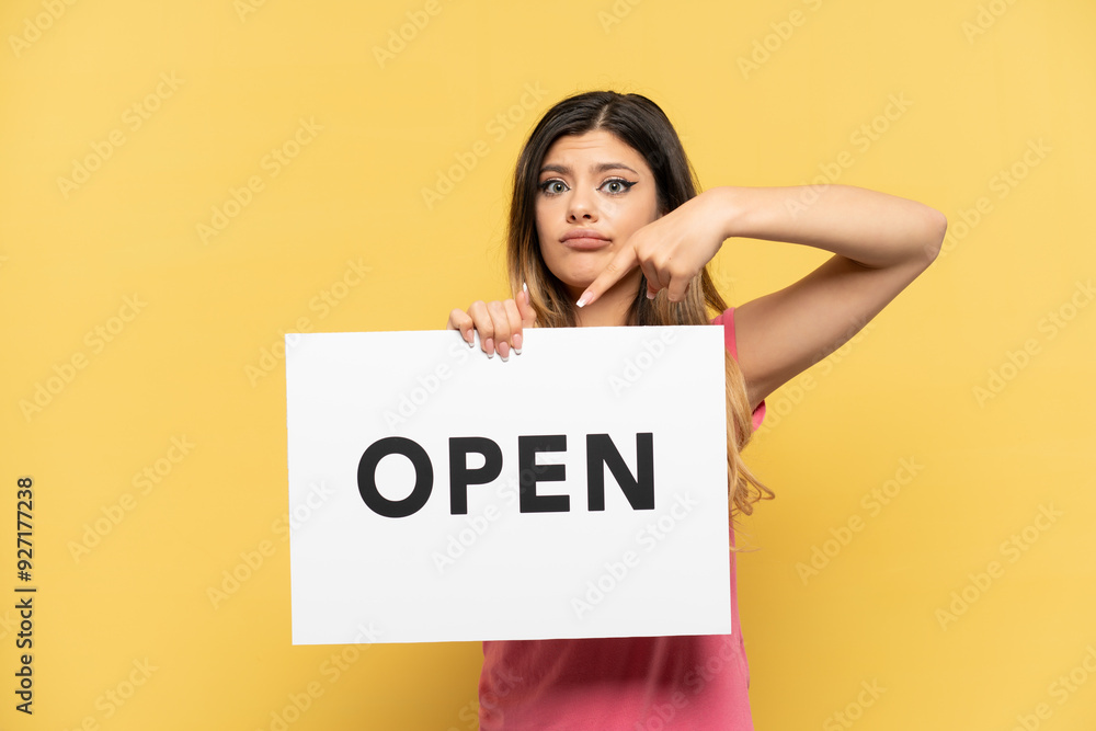 Young Russian girl isolated on yellow background holding a placard with text OPEN and pointing it Young Russian girl isolated on yellow background holding a placard with text OPEN and pointing it