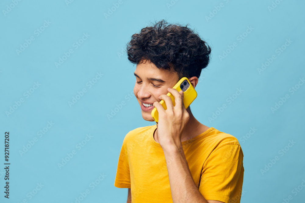 Young man speaking on a yellow smartphone, smiling happily, wearing a bright yellow t shirt against a solid blue background Joyful expression, casual vibe, modern technology Young man speaking on a yellow smartphone, smiling happily, wearing a bright yellow t shirt against a solid blue background Joyful expression, casual vibe, modern technology