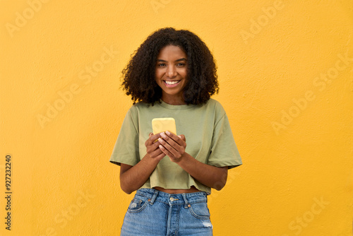 Young happy African gen z teen girl standing at yellow wall looking at camera holding smartphone. Smiling Black woman using mobile phone advertising summer shopping sale or mobile apps. Portrait. photo