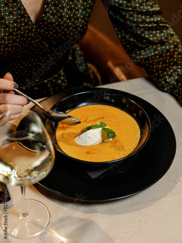 cute girl in a beautiful dress is eating a delicious cream soup with cream cheese, fresh vegetables, and fragrant herbs, served on a white plate in a cozy, charming restaurant