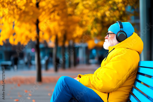 Elderly man in yellow jacket and blue beanie enjoying music with headphones on a bench during autumn, surrounded by vibrant yellow foliage. photo