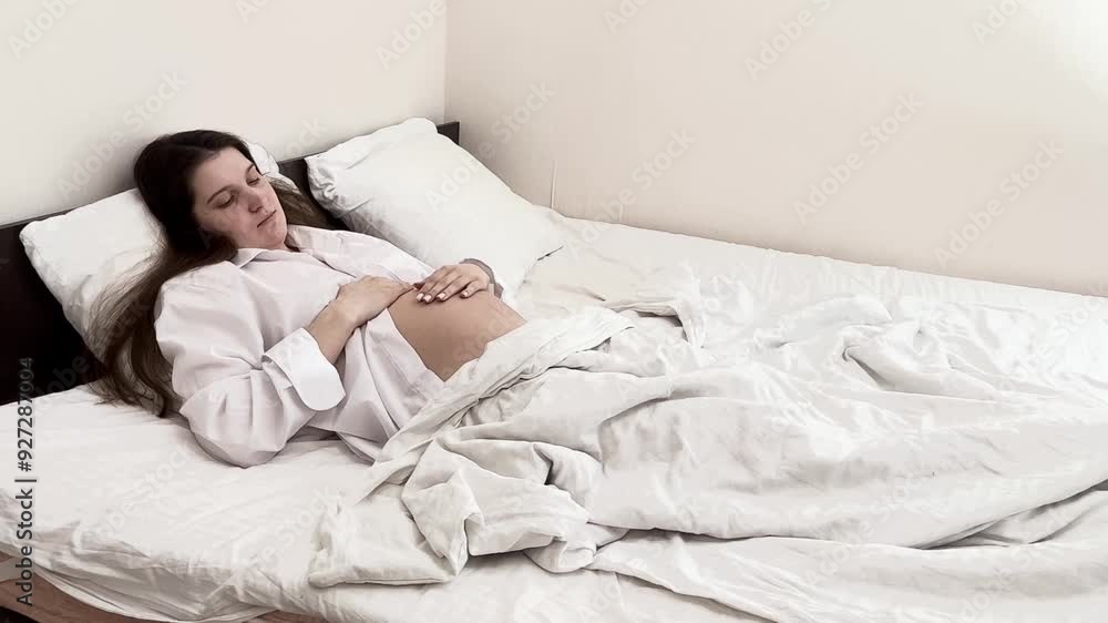 Pregnant girl with dark hair on the bed in a white shirt. International Day of the Midwife. Mothers Day. International Day of Families. Lazy Mom Day Pregnant girl with dark hair on the bed in a white shirt. International Day of the Midwife. Mothers Day. International Day of Families. Lazy Mom Day