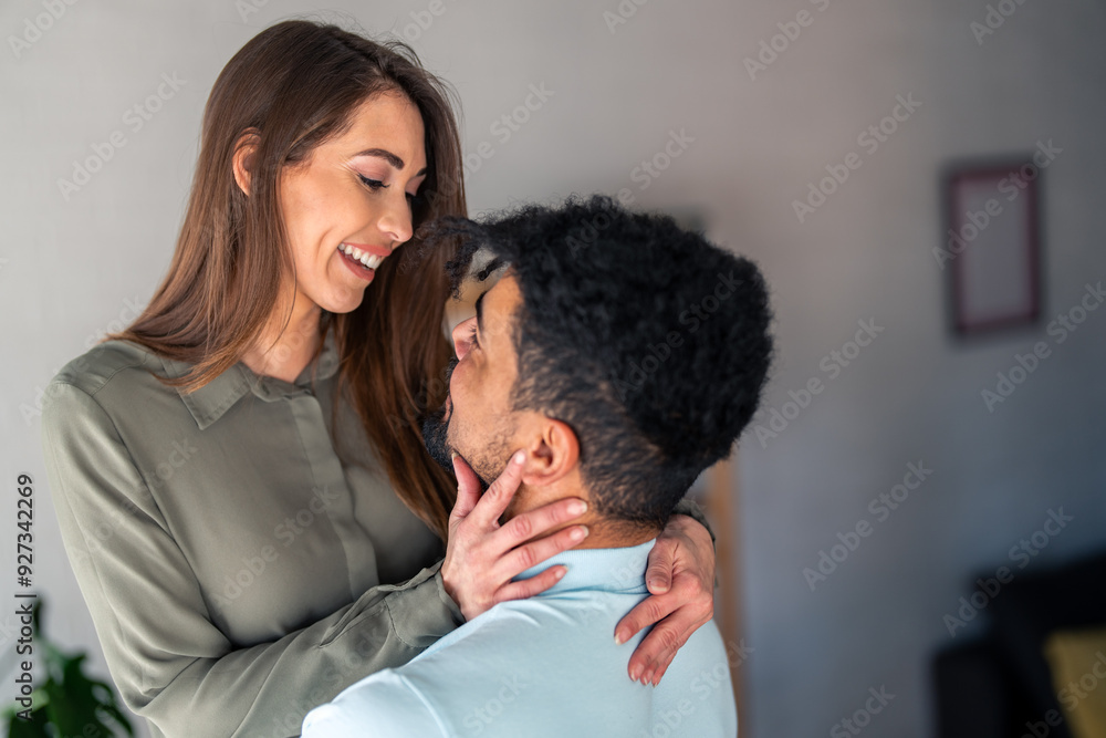 A smiling Caucasian woman lovingly wraps her arms around her mixed-race partner, both dressed casually, sharing an intimate moment within a home setting. A smiling Caucasian woman lovingly wraps her arms around her mixed-race partner, both dressed casually, sharing an intimate moment within a home setting.