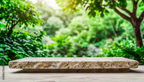 An empty stone table top centered on a white surface, set against a blurred background of vibrant green foliage. The scene is designed for displaying products in a natural, serene setting.