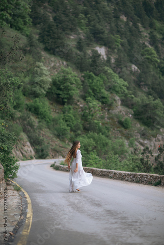 A woman in a white dress is walking down a road. The road is surrounded by trees and the woman is looking ahead.