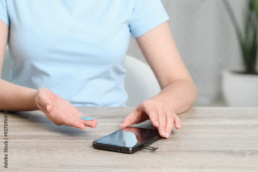Fototapeta premium Woman holding SIM card near smartphone at wooden table indoors, closeup