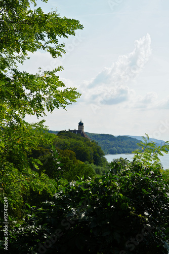 Medieval castle perched on a hill by a river, under a blue sky with clouds, surrounded by lush forest