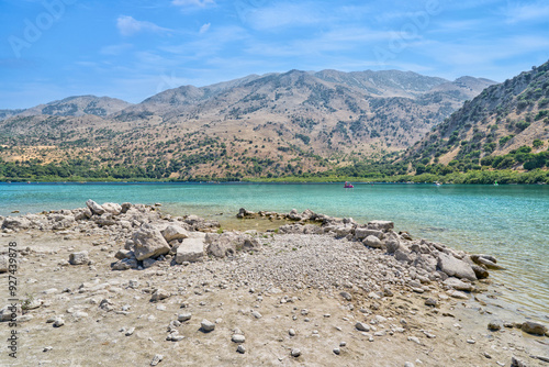 Scenic landscape of stony shoreline of Mediterranean Kournas Lake - Crete, Greece. Beautiful blue sky, mountains in the background, calm azure water. photo