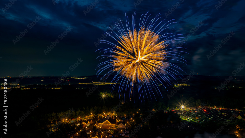 Golden And Blue Fireworks Burst Over A Well-Lit Estate, Surrounded By Trees And Fields, With Distant City Lights Adding Depth To The Festive Nighttime Scene. Golden And Blue Fireworks Burst Over A Well-Lit Estate, Surrounded By Trees And Fields, With Distant City Lights Adding Depth To The Festive Nighttime Scene.